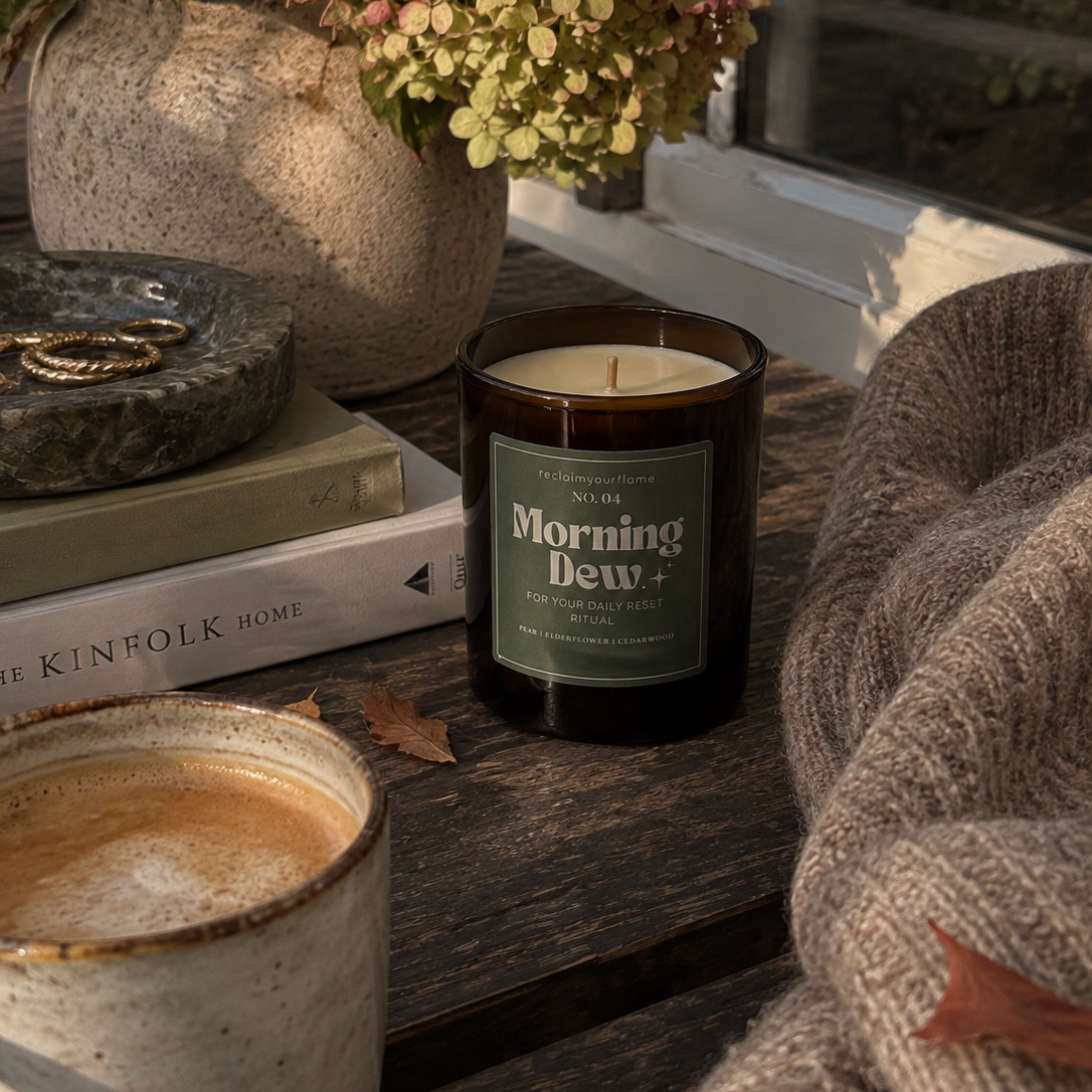 A dark amber glass candle jar labeled 'Morning Dew' sits on a rustic wooden table beside a mug of coffee, stacked books, a stone dish with rings, and a knitted sweater draped in warm sunlight near a window.