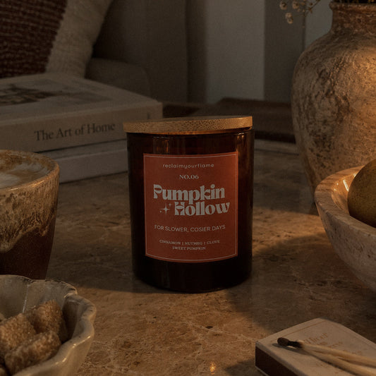 A dark amber glass jar candle with a light wood lid sits on a stone tabletop in a cozy indoor setting. The front label reads 'Pumpkin Hollow''.