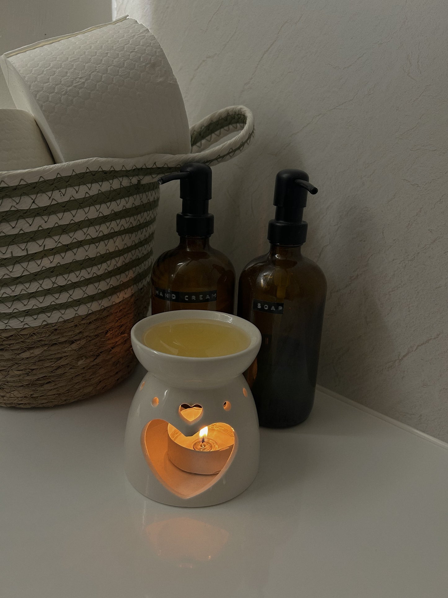A small white wax warmer with a lit candle inside on a white surface, with two brown bottles and a woven basket in the background.