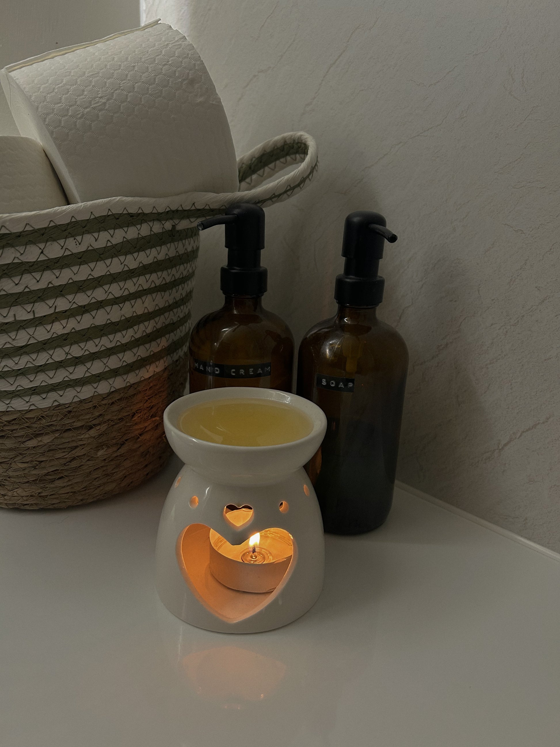 A small white wax warmer with a lit candle inside on a white surface, with two brown bottles and a woven basket in the background.