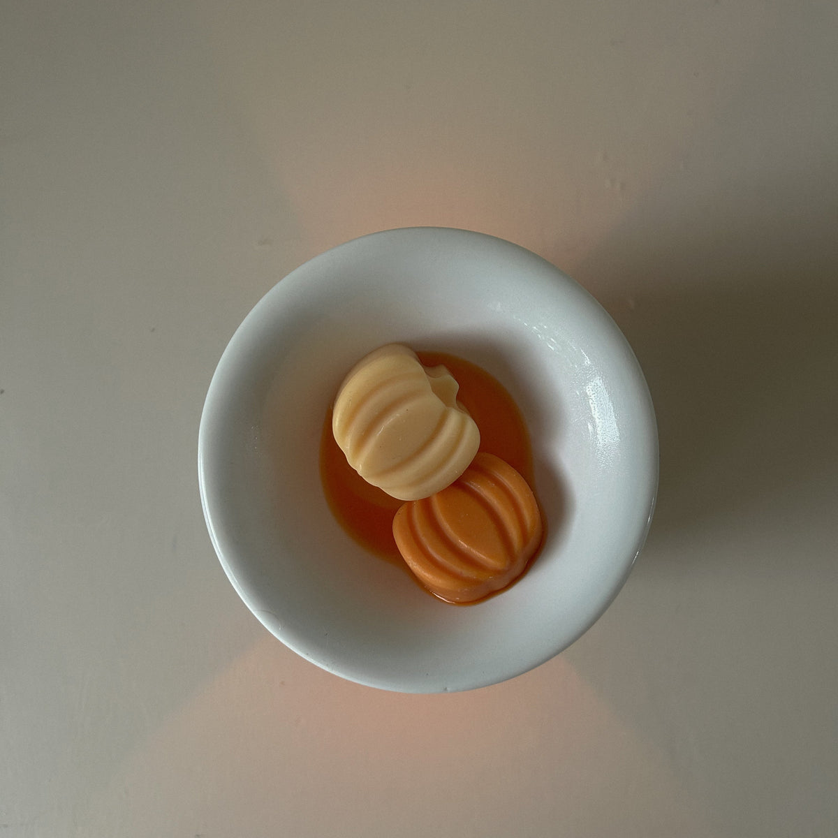Two small decorative items, one cream-colored and one orange, resting in a small white ceramic bowl on a plain beige surface.