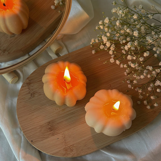 Two pumpkin-shaped candles with warm orange glow, displayed on a wooden board with dried baby's breath flowers and draped white fabric.
