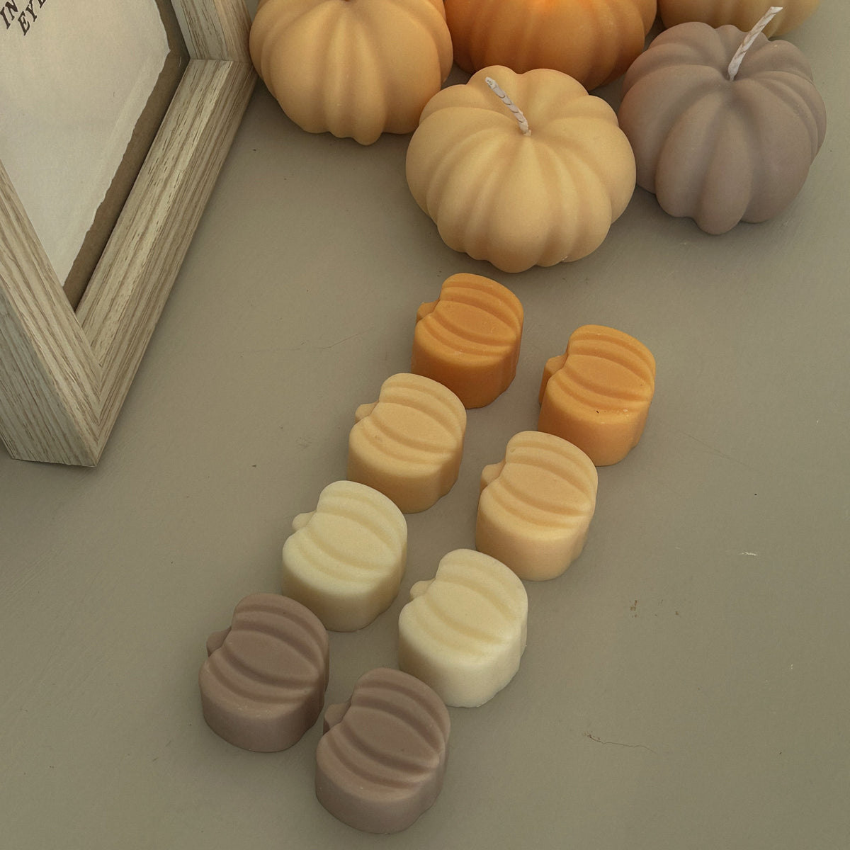 Pumpkin-shaped candles in warm autumn shades, including unlit beige, orange, and taupe pumpkin forms, with one orange candle lit and additional pumpkin wax pieces arranged in front on a neutral surface.