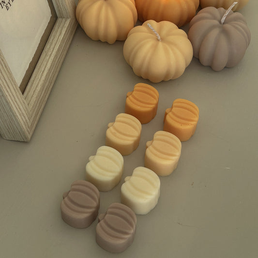 Pumpkin-shaped candles in warm autumn shades, including unlit beige, orange, and taupe pumpkin forms, with one orange candle lit and additional pumpkin wax pieces arranged in front on a neutral surface.