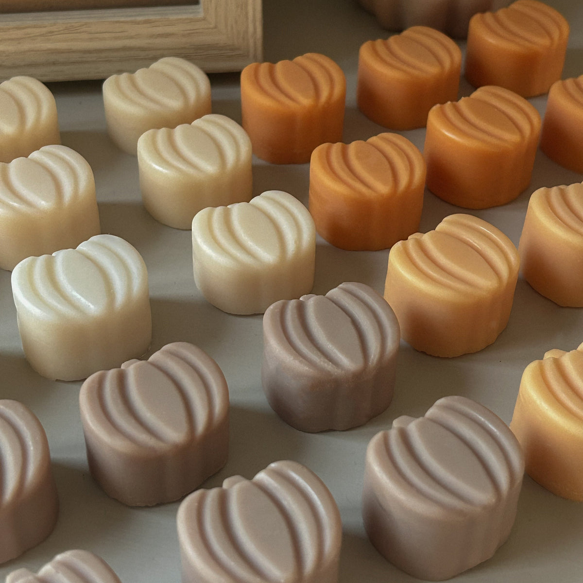A close-up arrangement of small, ribbed, pumpkin-shaped wax melts or soaps in warm neutral tones including cream, tan, peach, and taupe, displayed on a tabletop with a framed picture and a decorative pumpkin in the background.