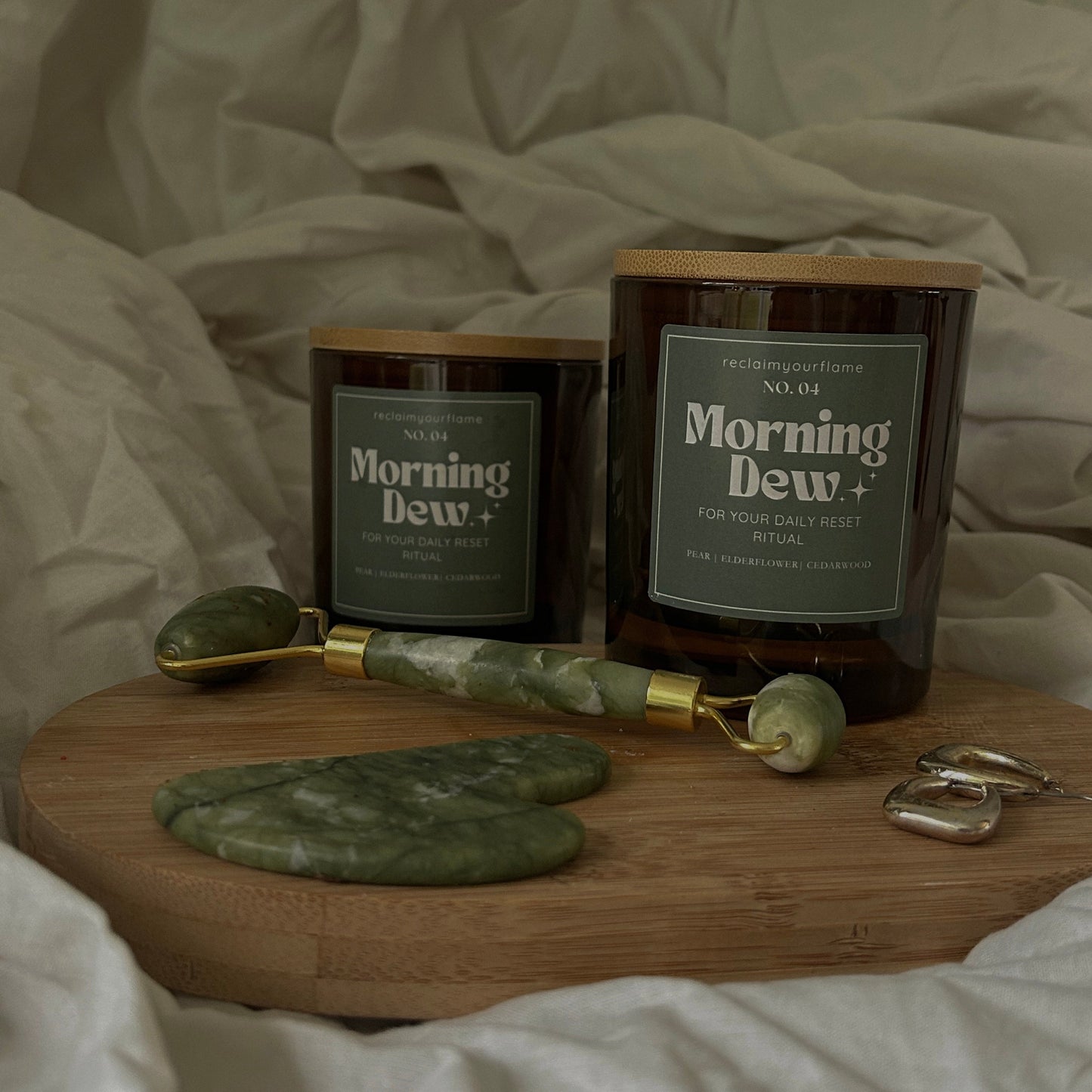Two 'Morning Dew' candles on a wooden tray with a green gua sha and a jade roller on a white fabric background.