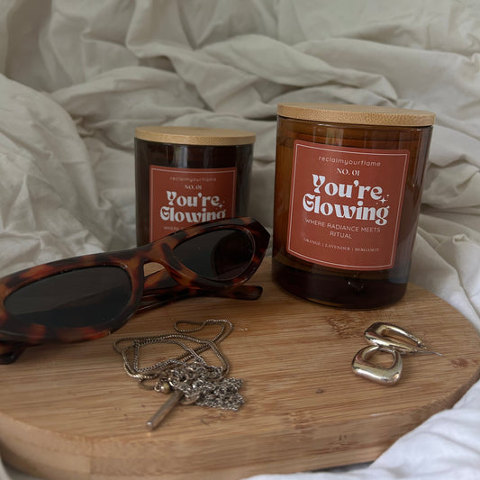 Two candles  with 'You're Glowing' on the label on a wooden board with sunglasses and jewellery.