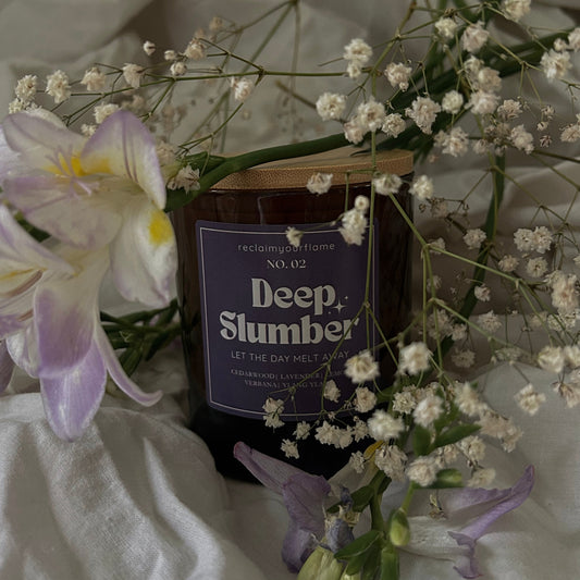 Candle labeled 'Deep Slumber' surrounded by flowers on a white fabric background