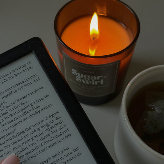 A sugar swirl candle lit in the background of someone reading a book with a hot drink.