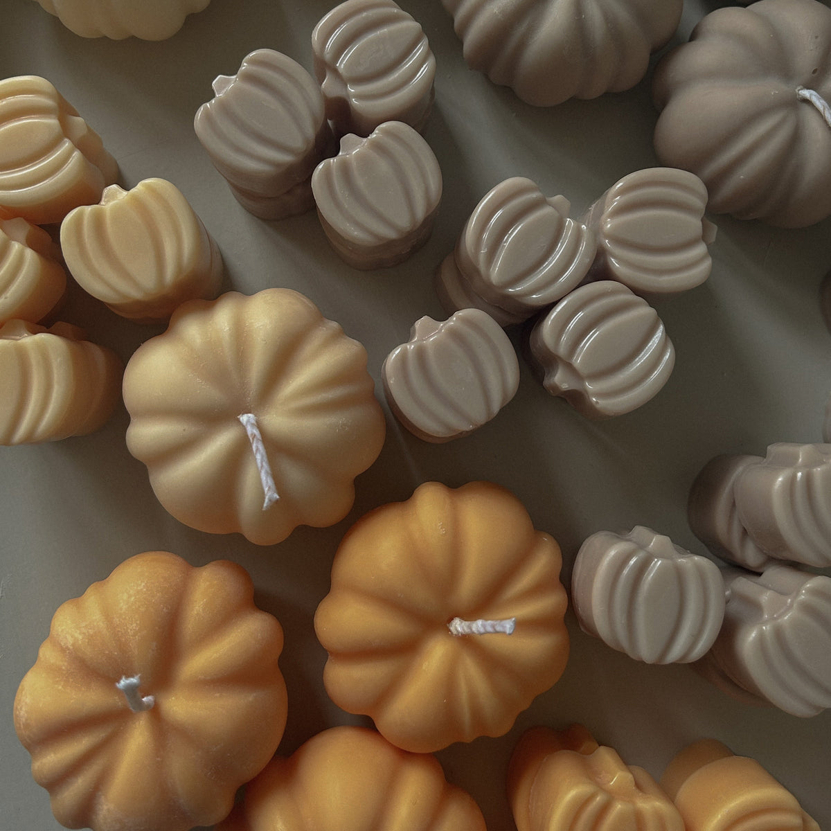 A collection of pumpkin-shaped candles in warm autumn shades, including cream, taupe, peach, and orange, arranged closely together on a neutral surface.
