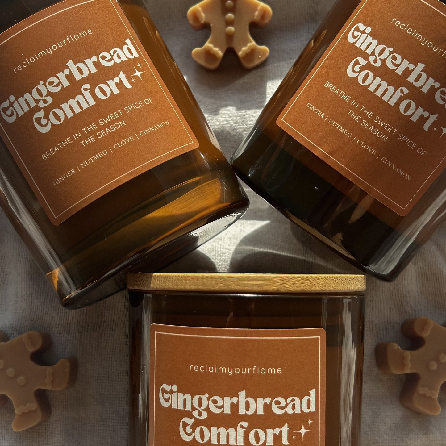 Three container candles with 'Gingerbread Comfort' on the label on a bedspread with mini gingerbread surrounding.